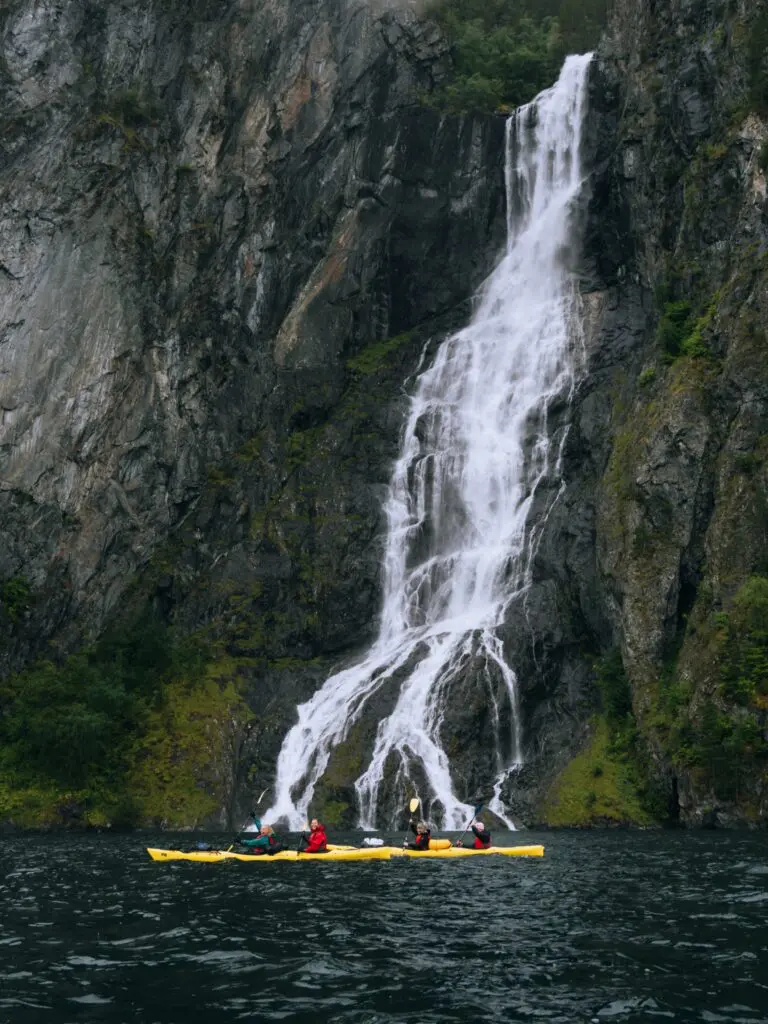 kajakfahrende Personen paddeln an einem großen Wasserfall in einem Fjord vorbei