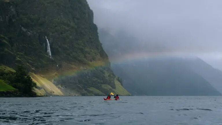 Zwei Menschen fahren auf einem Fjord mit einem Regenbogen und einem Wasserfall Kajak