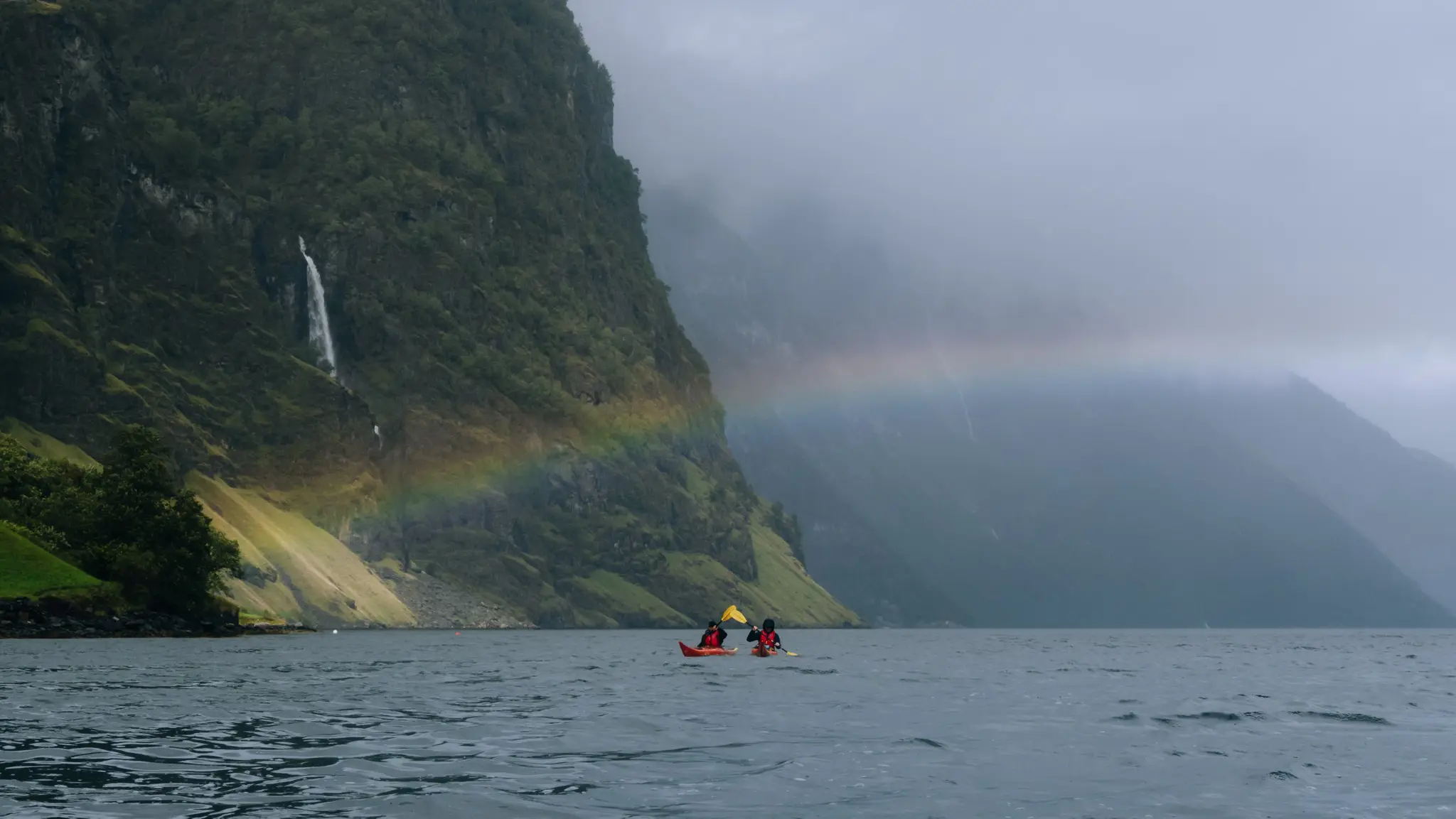 Zwei Menschen fahren auf einem Fjord mit einem Regenbogen und einem Wasserfall Kajak