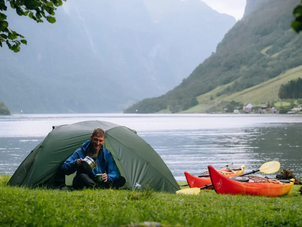 Mann, der draußen vor einem Zelt am Fjord Kaffee einschenkt, mit Kajaks Grammatikkorrigiert: Mann, der draußen vor einem Zelt am Fjord Kaffee einschenkt, mit Kajaks.