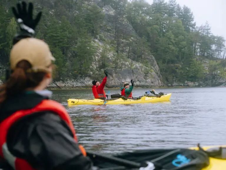 Drei Personen paddeln mit Kajaks auf einem See, umgeben von bewaldeten Felsklippen.