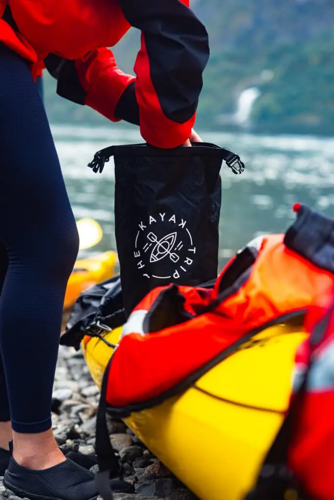 Person, die an einem felsigen Ufer neben einem Kajak einen Packsack packt