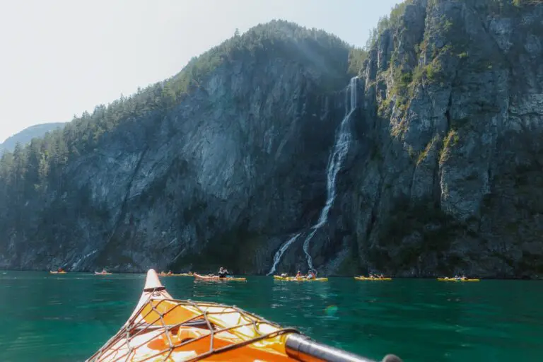 kajakfahrende Personen paddeln an einem großen Wasserfall in einem Fjord vorbei